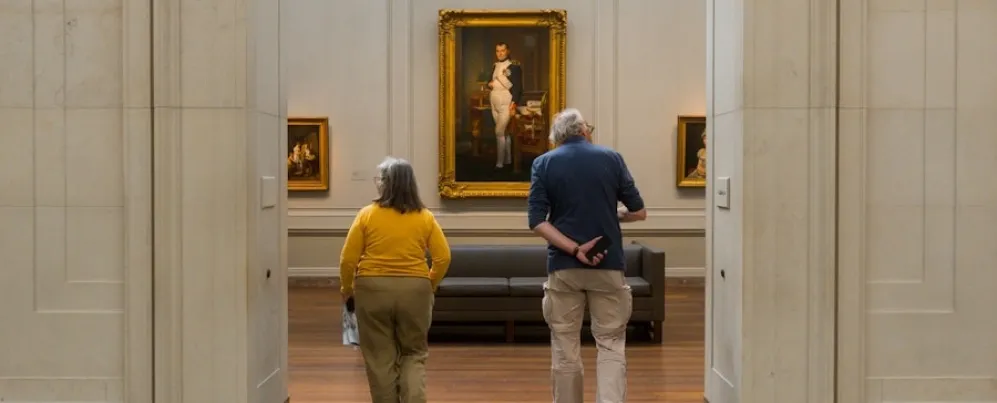 Two people walk into a museum gallery toward a large framed portrait hanging on the far wall.
